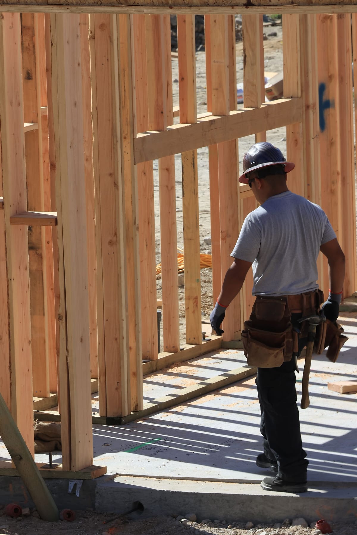 A framer in a hard hat and tool belt stands beside a wall of fresh studs.